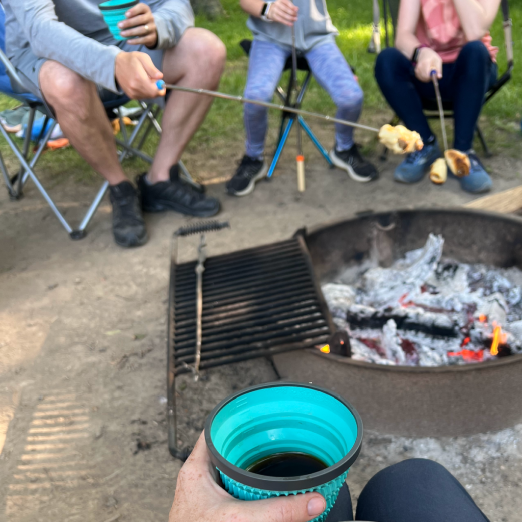 hand holding camp mug of coffee next to campfire with family roasting marshmallows behind