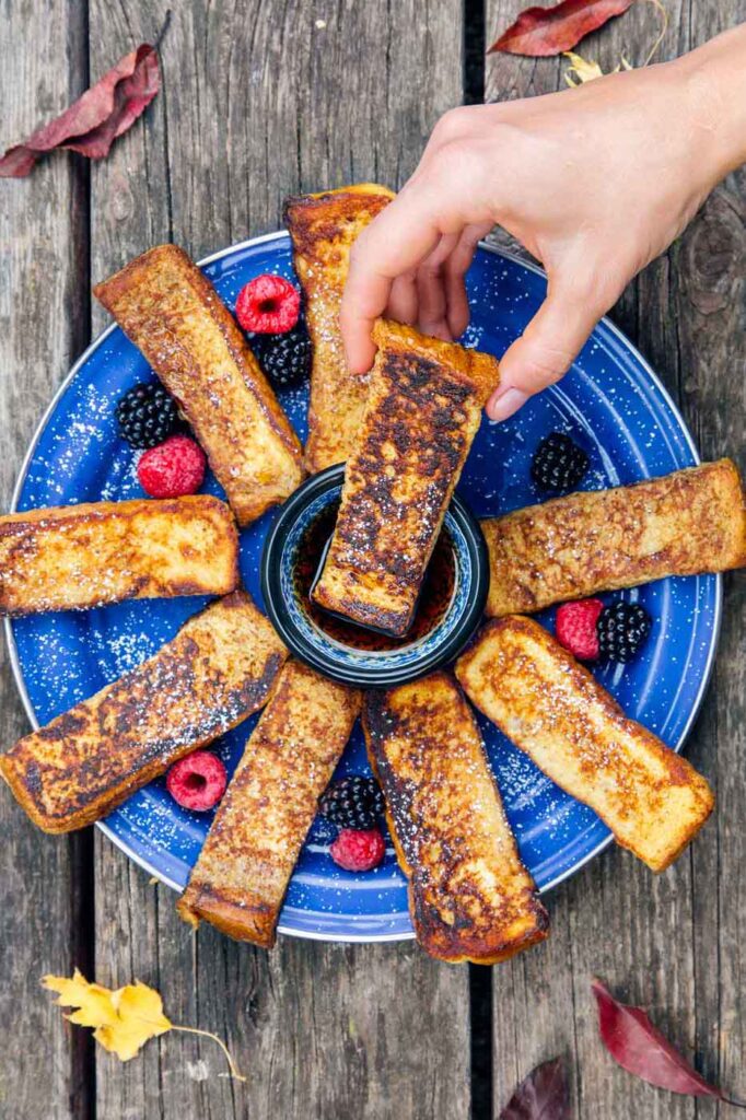 french toast sticks on a blue plate with berries, with a hand dipping one into a bowl of syrup