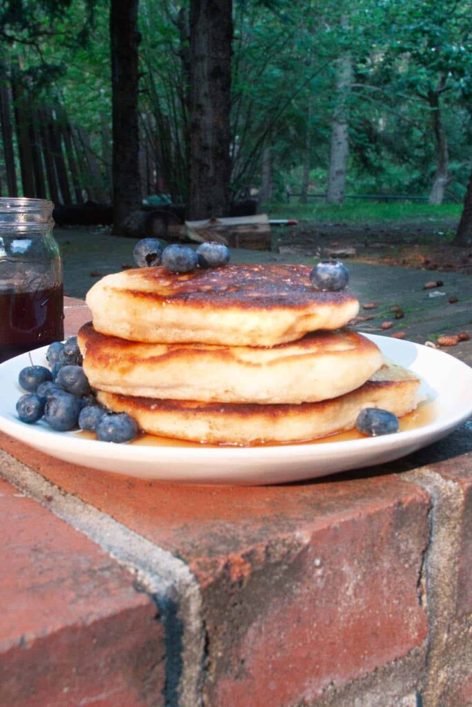 three pancakes stacked on a plate with blueberries, with the woods behind